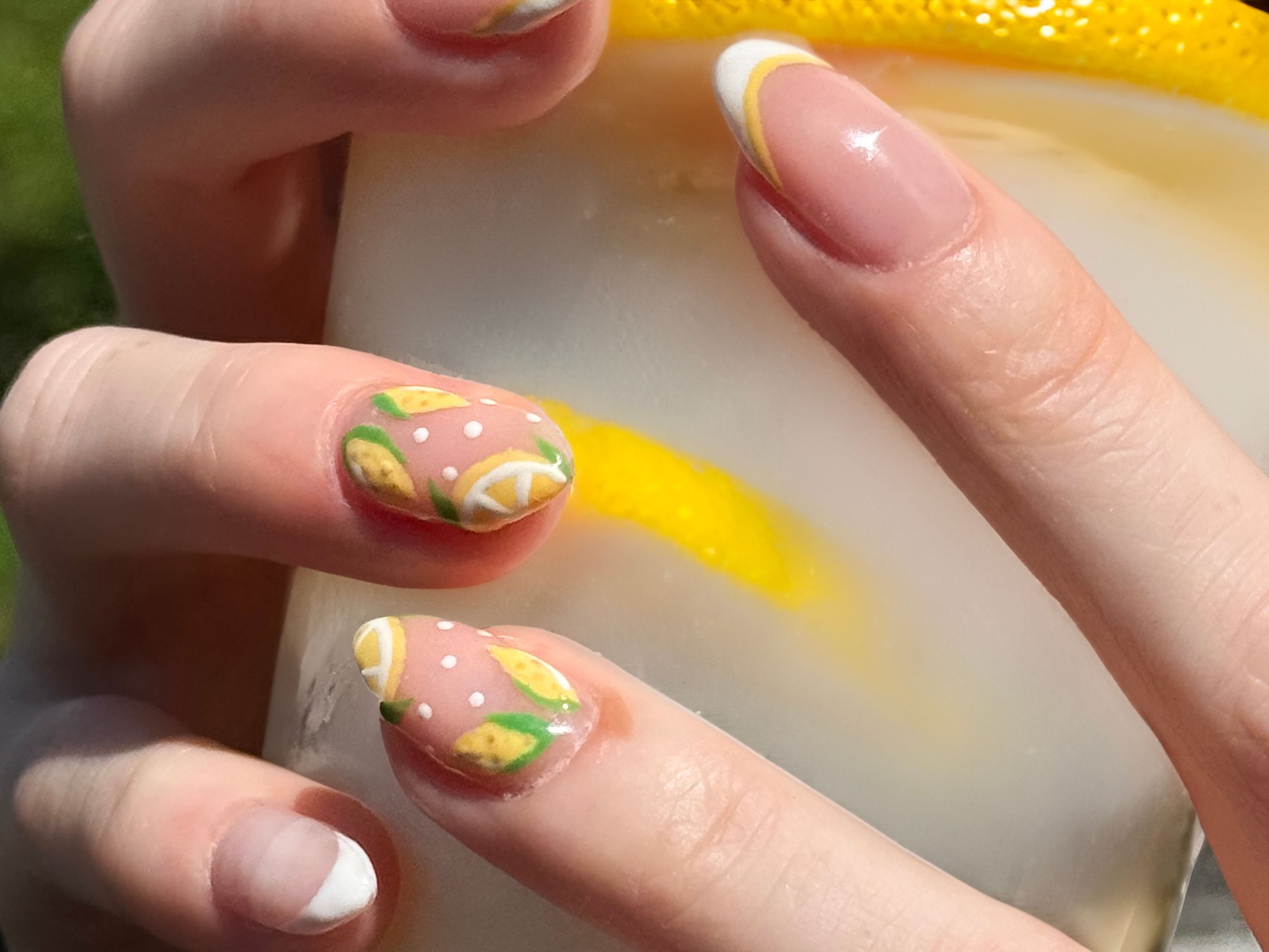 Full frame image of unrecognisable woman's hands holding cloudy, old fashioned, lemonade drink in plastic cup, showing nail varnish manicure design of lemon fruit and citrus slices, sunny outdoor environment background, focus on foreground